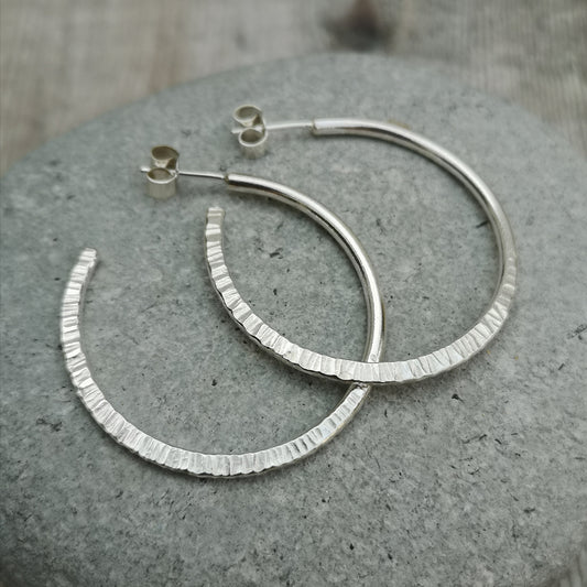 Close-up of large handmade sterling silver hoop earrings with a unique hammered texture, displayed on a smooth grey stone background.