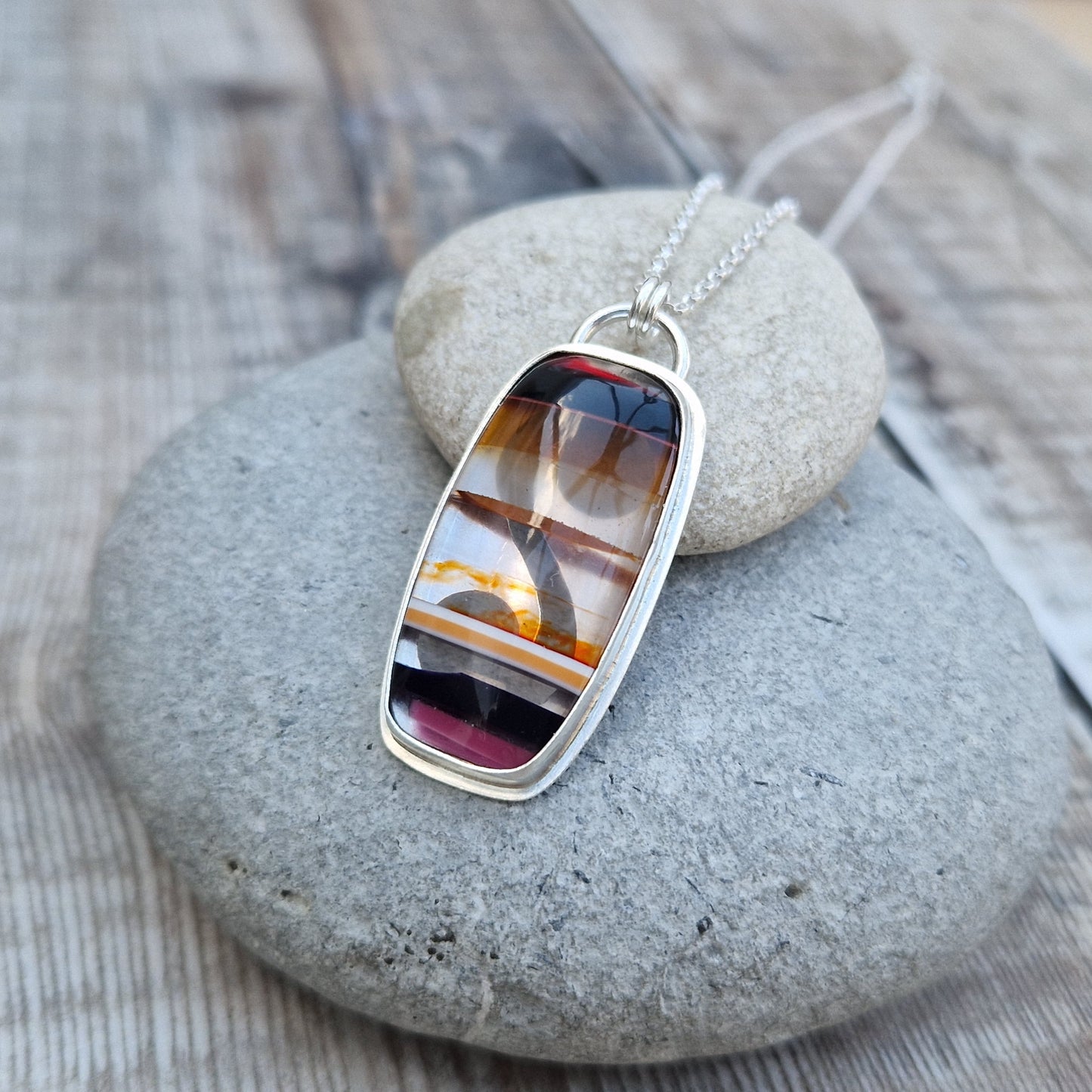 Pendant is a rectangle cut Surfite stone set in a Sterling Silver bezel surround, suspended from a silver chain via two small circle links. The coloured horizontal surfite stripes are black, dark brown, opaque, brown, orange flecks, black dark pink.