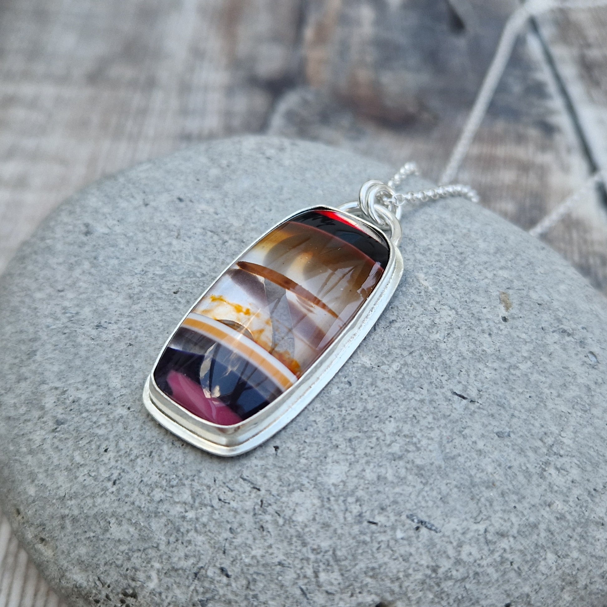 Pendant is a rectangle cut Surfite stone set in a Sterling Silver bezel surround, suspended from a silver chain via two small circle links. The coloured horizontal surfite stripes are black, dark brown, opaque, brown, orange flecks, black dark pink.