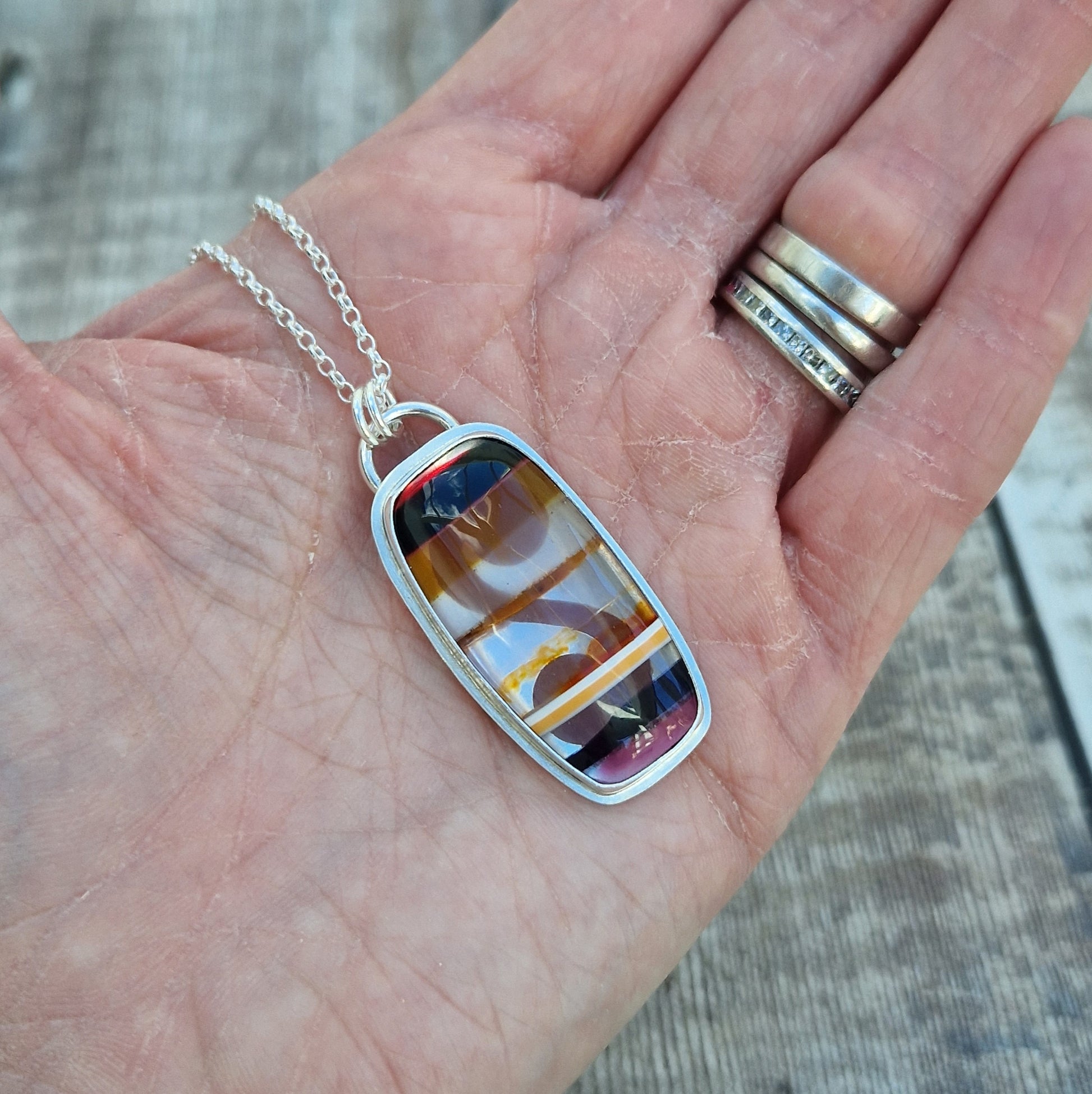 Pendant is a rectangle cut Surfite stone set in a Sterling Silver bezel surround, suspended from a silver chain via two small circle links. The coloured horizontal surfite stripes are black, dark brown, opaque, brown, orange flecks, black dark pink.