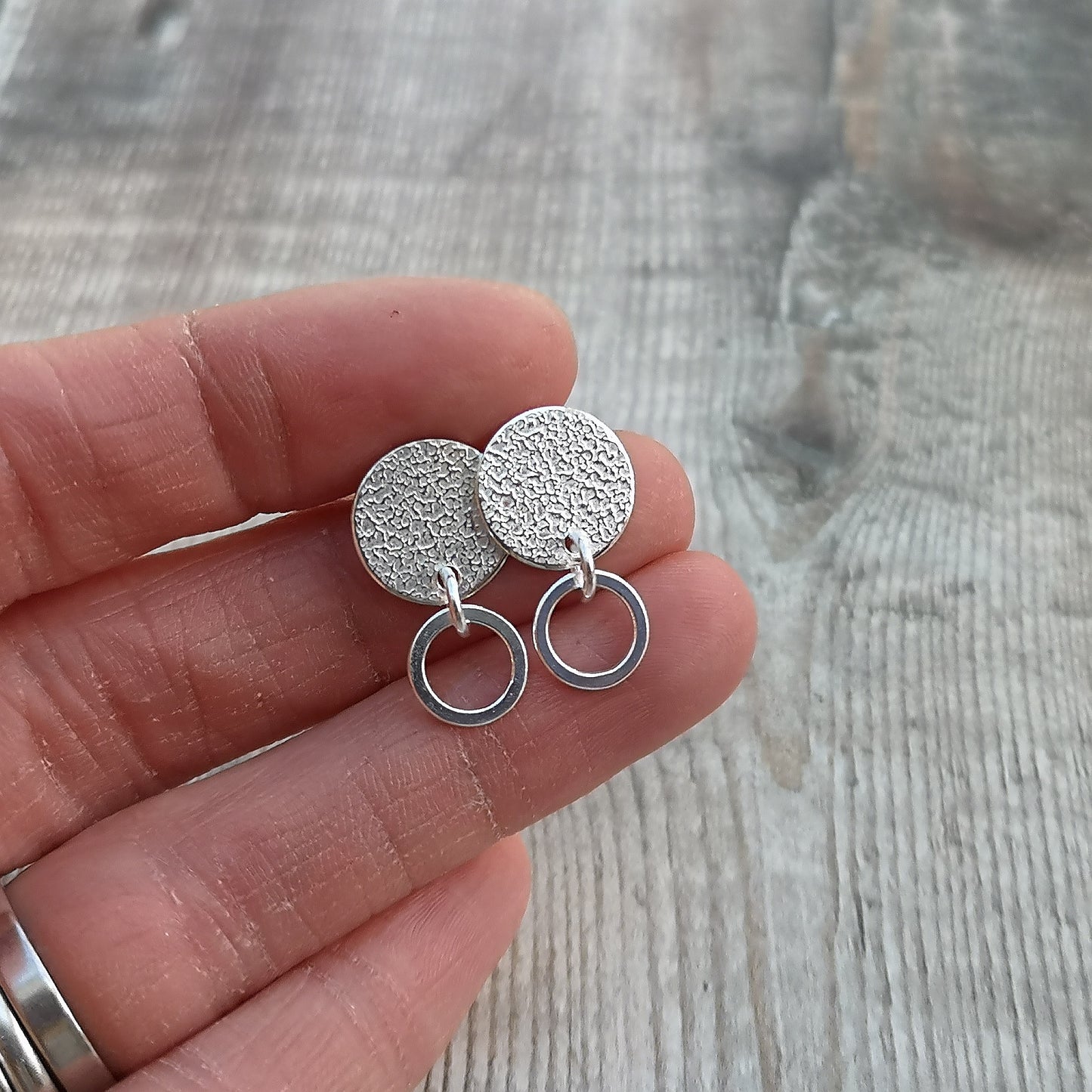 Close-up of a hand holding a pair of handmade stud earrings with a hammered silver disc and a small hoop, placed against a light wooden background with soft-focus dried flowers in the background.