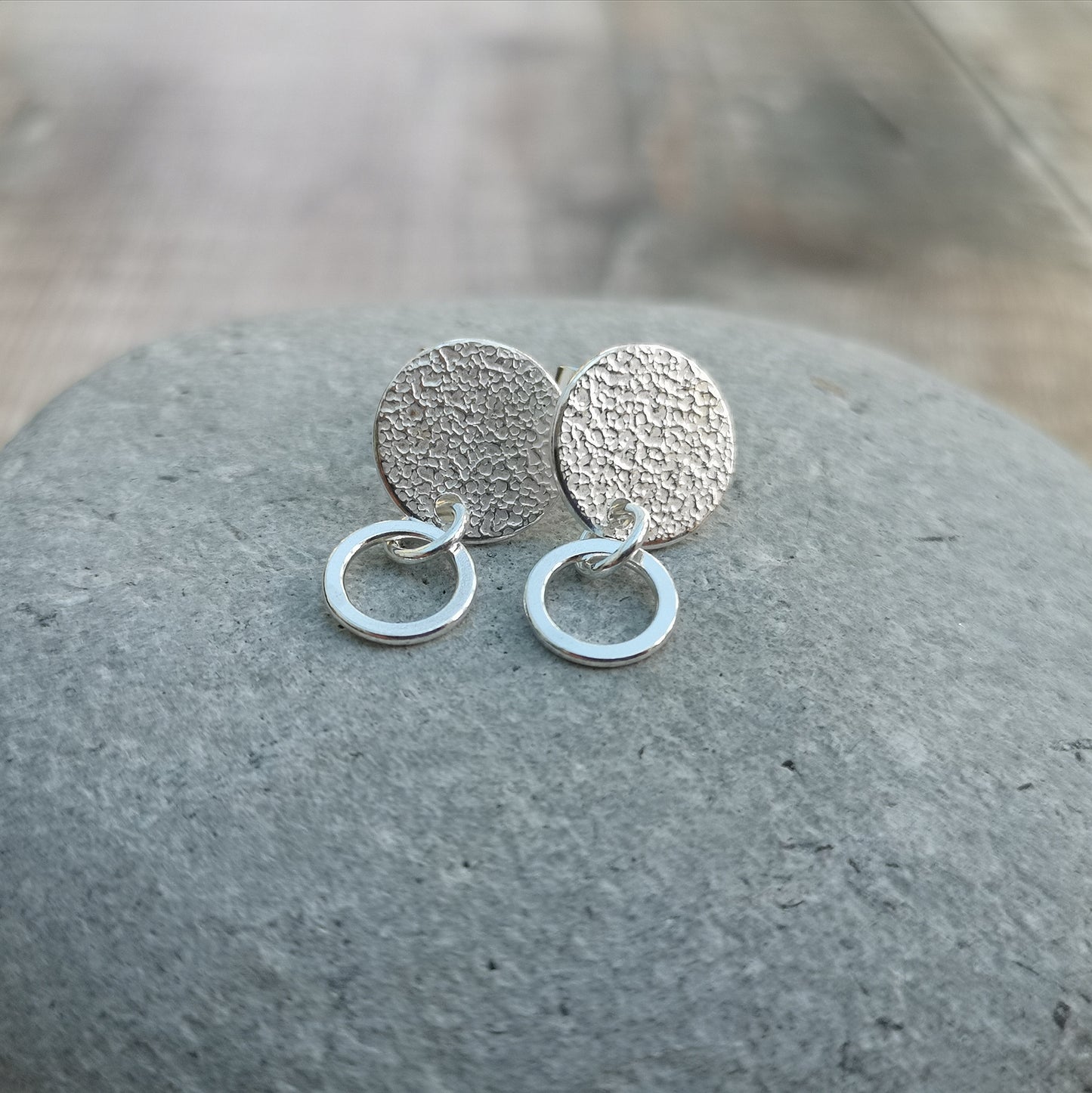 A pair of handmade stud earrings featuring a hammered silver disc with a small hoop, displayed on a smooth grey stone with purple and beige dried flowers softly blurred in the background.